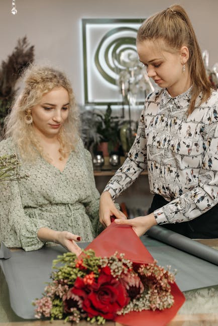 A professional florist from floristcroftonpark.co.uk, wearing a patterned blouse, is carefully wrapping a vibrant bouquet featuring red roses, pink and white flowers, and greenery in red wrapping pape