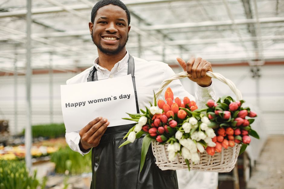 A male florist with a warm smile holding a white wicker basket filled with fresh, vibrant tulips in shades of pink, orange, and white, arranged with green foliage. He is standing inside a greenhouse w