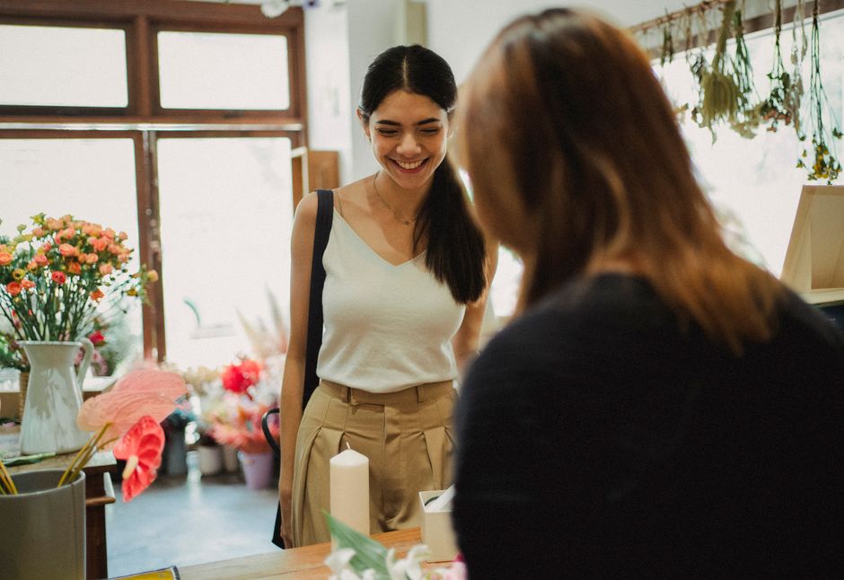 A young woman with dark hair smiling as she receives a floral gift from a florist inside a flower shop. The bouquet features a variety of flowers, including pink anthuriums, orange and red roses, and 