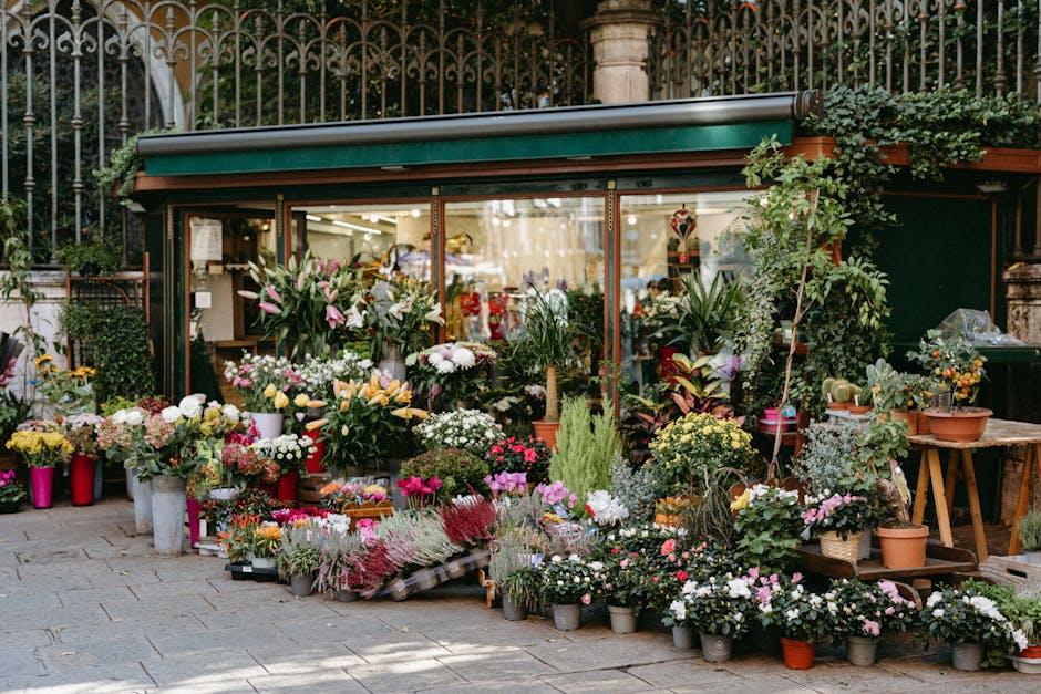 An outdoor flower shop displays a wide variety of fresh floral arrangements and potted plants arranged on the pavement in front of a glass storefront, with lush greenery and decorative plants surround