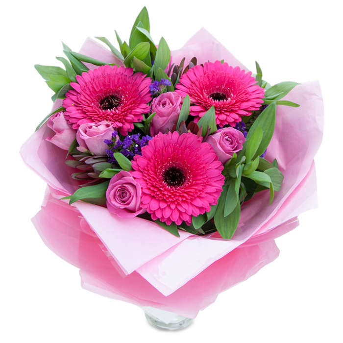 I see a bouquet that balances playful colour with gentle refinement: three fuchsia Gerbera daisies burst forward with graphic, petal-rich faces and deep centres that create a striking focal point, while surrounding lavender-pink roses curl open in layered, velvety spirals, their dusty tones tempering the brightness. Scattered among them are small clusters of purple statice, adding a textured, almost wildflower note and a cool violet counterpoint to the pinks. The foliage-broad, glossy salal leaves and a few silver-green eucalyptus accents-wraps the blooms in a lush, calming green, offering both structure and the faint, balsamic lift of eucalyptus scent. The bouquet is wrapped for gifting: a pale pink matte tissue cradles the stems and petals, and an outer sheet of slightly crinkled, translucent fuchsia-pink cellophane gives the package a subtle lustre; the careful folds and neat ties reflect the maker's attention to presentation. At the base the clear rim of a glass vase peeks into view, signaling that the arrangement is ready to be watered and displayed immediately. The overall impression is one of warmth and thoughtful cheer-an arrangement that would brighten a bedside table, bring colour to a living room, or arrive as a charming surprise from a neighbour in Crofton Park or a nearby walk through Blythe Hill Fields. It feels like a personal celebration in bloom, suited to birthdays, Mother's Day, or an intimate anniversary.